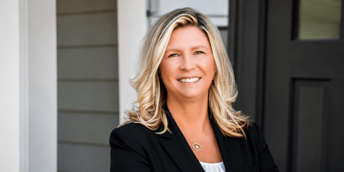 Smiling woman with blonde hair wearing a black blazer standing outside a house