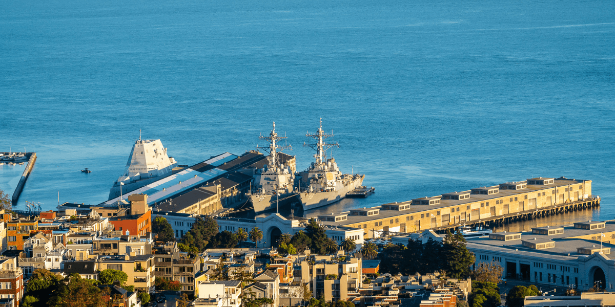 Two military ships docked at a harbor with city buildings in the foreground