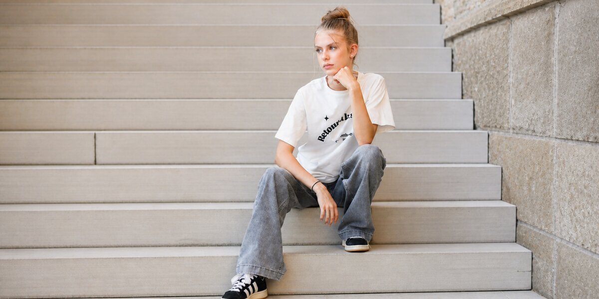 Young woman with blonde hair in a bun sits pensively on gray steps. She's wearing a white graphic tee, loose jeans, and black sneakers.