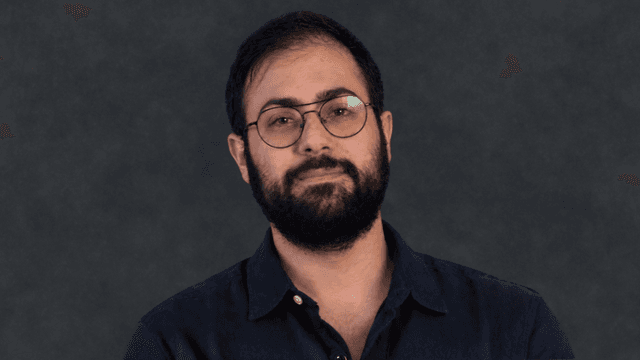 A man with glasses and a beard poses confidently against a dark background