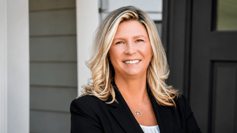 Smiling woman with blonde hair wearing a black blazer standing outside a house