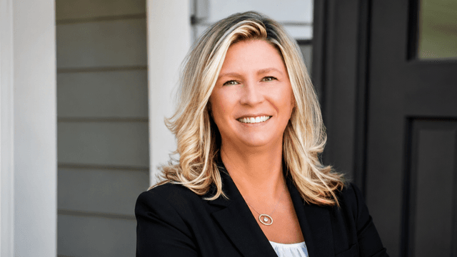 Smiling woman with blonde hair wearing a black blazer standing outside a house