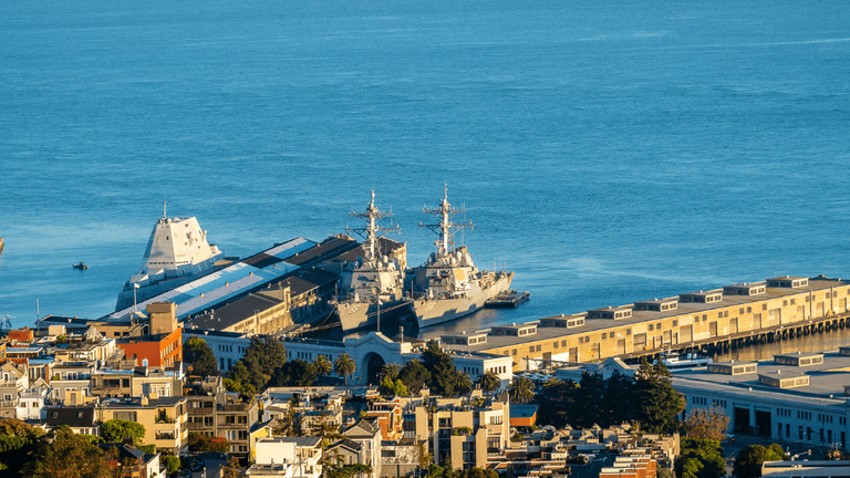 Two military ships docked at a harbor with city buildings in the foreground