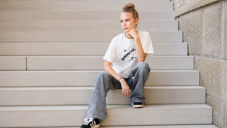 Young woman with blonde hair in a bun sits pensively on gray steps. She's wearing a white graphic tee, loose jeans, and black sneakers.