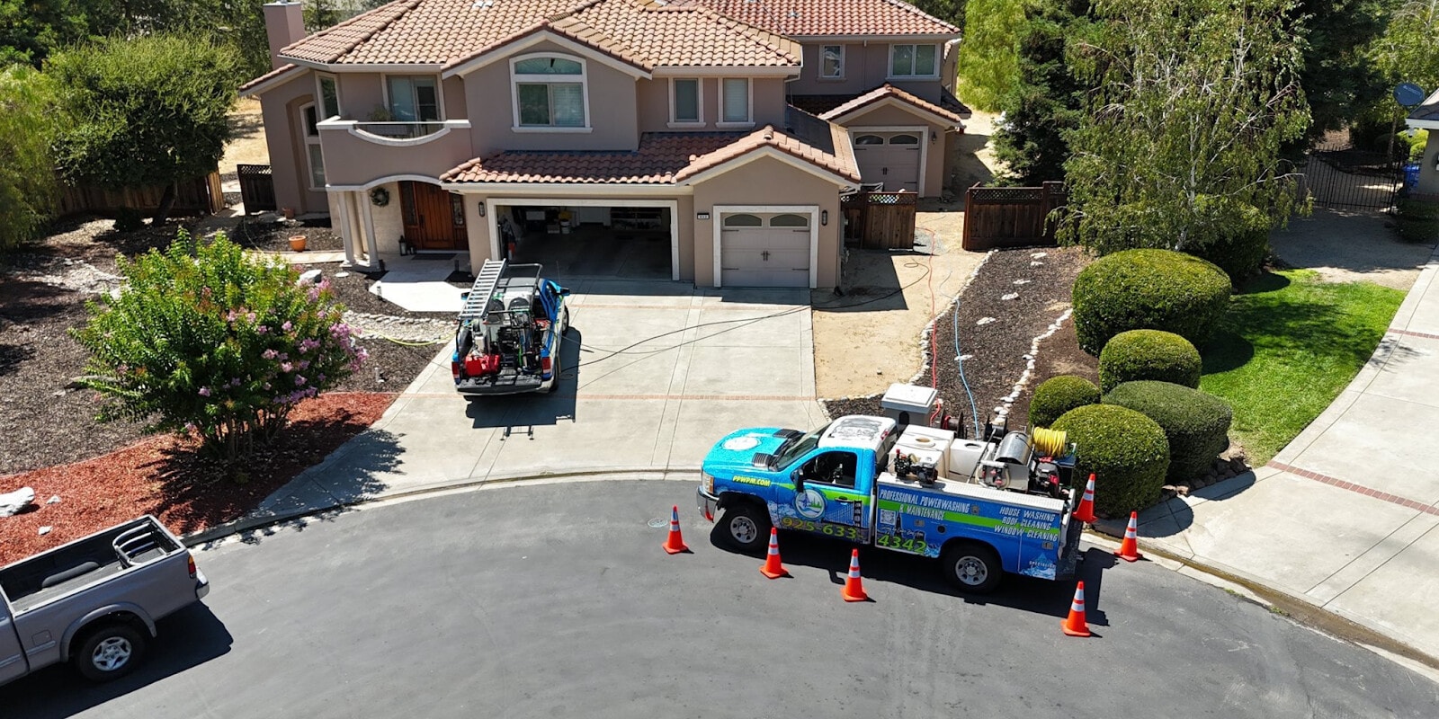 Blue truck with power washing equipment parked outside a residential property, surrounded by traffic cones.