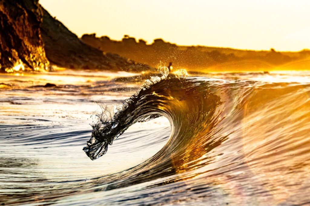 Golden sunrise illuminates a curling ocean wave captured from inside the surf near a rocky coastline.